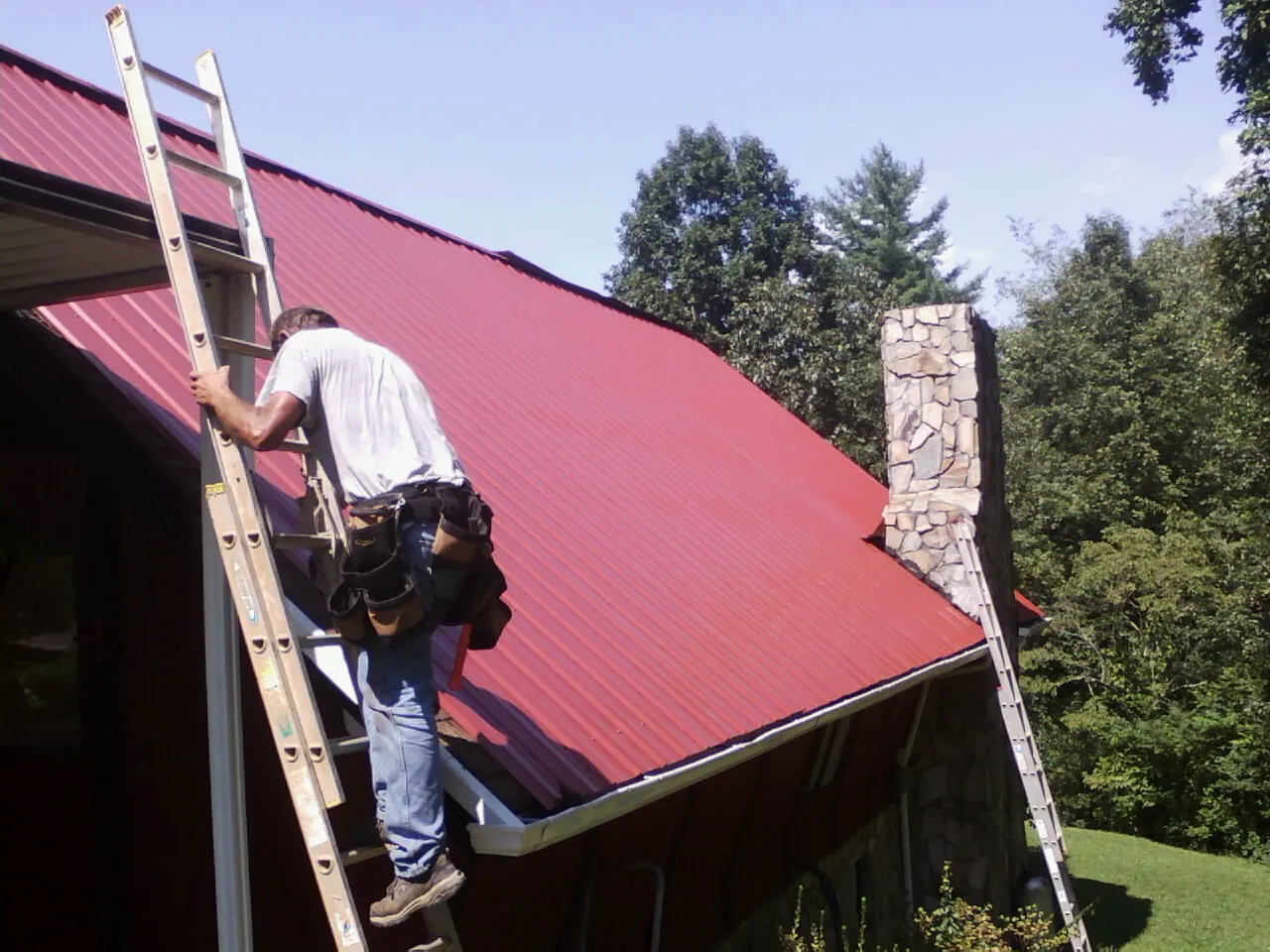 Roofer on a ladder beside a red metal roof and stone chimney — Soffit & Fascia Repair by Roofing Brothers in Bainbridge Island
